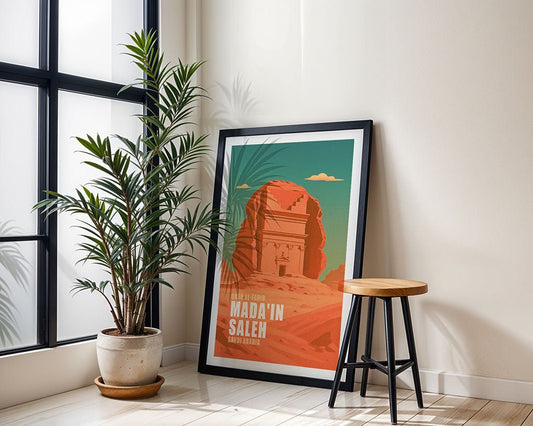 The FN Prints "Qasr al-Farid Madain Saleh" framed poster leans against a light wall beside a potted plant and wooden stool in a bright room with large windows.