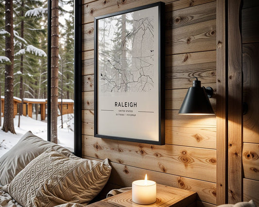 A cozy wooden cabin interior shows snow outside, featuring a minimalist aesthetic with an FN Prints Raleigh City Map above the bed adorned with patterned pillows. Nearby, a lit candle and black wall lamp on a table blend style and sustainability using eco-friendly inks.