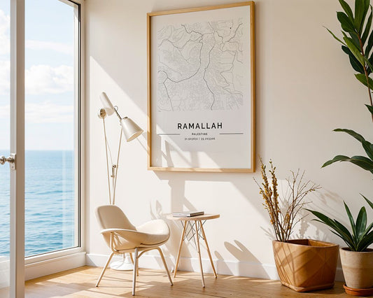 In a bright room, city wall art from FN Prints, featuring the Ramallah City Map, hangs above a minimalist chair and side table. A modern floor lamp stands nearby by large windows with ocean views. A corner potted plant adds greenery to the minimalist look.