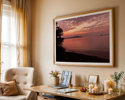 A home office with a wooden desk, laptop, candles, plants, and a beige chair. Above the desk hangs "Rose Gold Sunset" by FN Prints—a tranquil Goan beach sunset print. Sunlight filters through curtains, making the workspace peaceful.