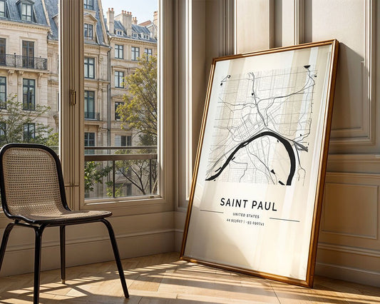 A FN Prints Saint Paul City Map in minimalist black and white, crafted on museum-quality paper, is framed and leans against a light wall in a sunlit room. A woven chair sits nearby while classic European-style buildings are visible through the window.