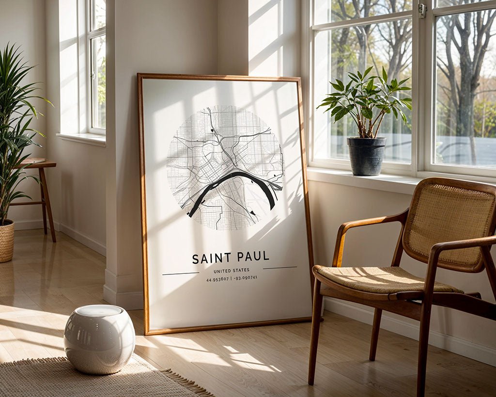 The FN Prints Saint Paul City Map, featuring a framed minimalist black and white design, leans against a well-lit wall. Sunlight floods the room with large windows highlighting a wooden chair, potted plants, and a small white pouf resting on the floor.