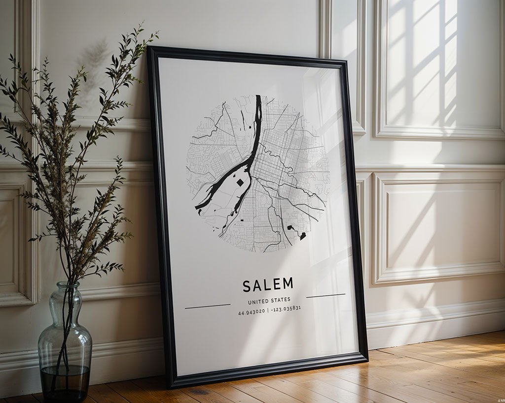 A framed black and white FN Prints' Salem City Map leans against a well-lit room's wall, exemplifying minimalist style. A tall plant in a glass vase stands to the left as sunlight filters through the window, casting subtle shadows on the wooden floor—perfect city wall art.