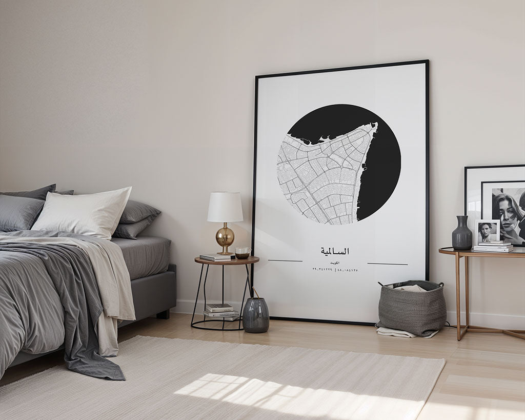 A modern bedroom features a bed with gray bedding, a wooden side table with a lamp and vase, and FN Prints' Salmiya City Map, printed on museum-quality fine art paper, leaning against the wall. A basket, rug, and framed photos adorn the hardwood floor.