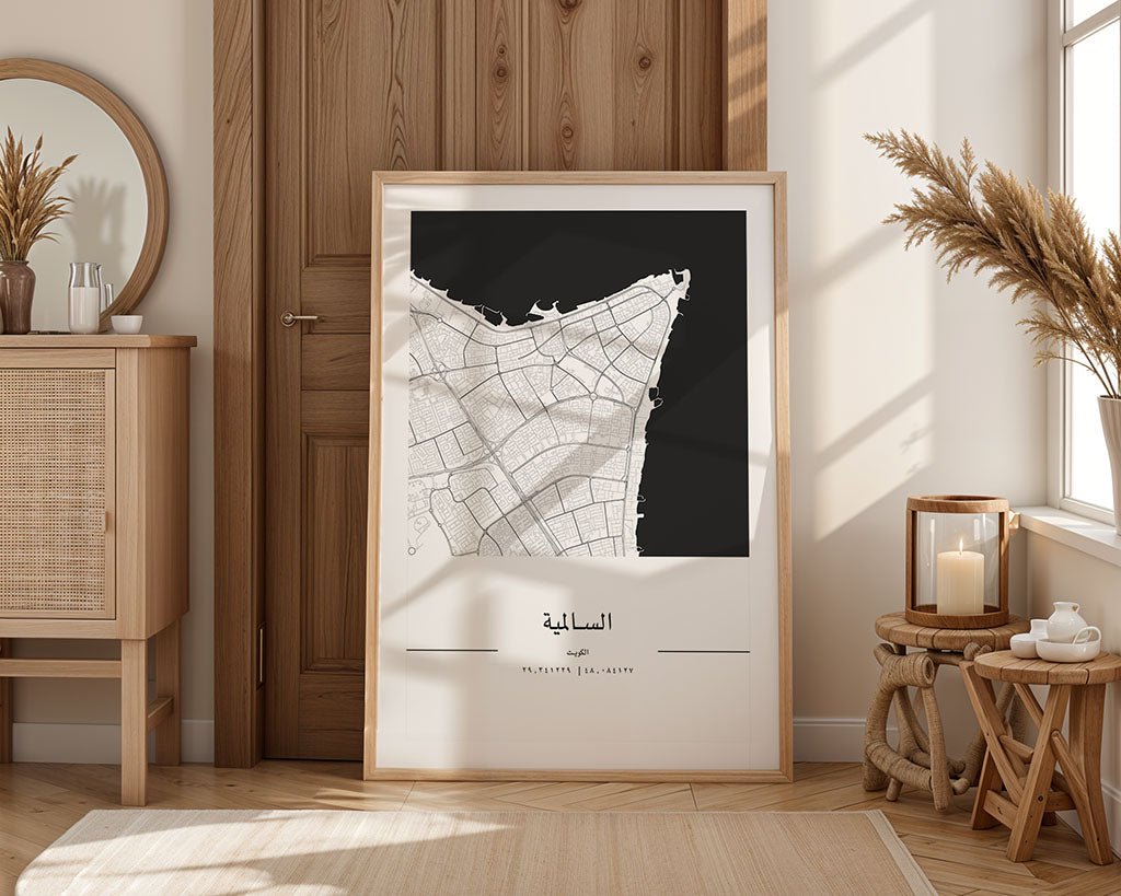 A framed FN Prints Salmiya City Map print leans against a wooden door in a sunlit room, accompanied by a wooden cabinet, wicker basket, round mirror, candles, and dried plants. This minimalist print on museum-quality fine art paper includes elegant Arabic text.