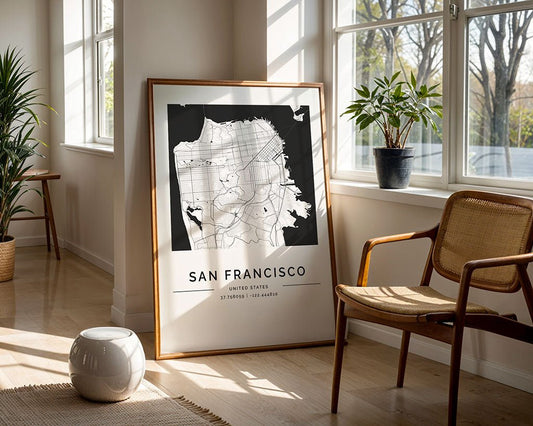The framed FN Prints' San Francisco City Map sits on the floor in a sunlit room, epitomizing modern interiors. Its monochrome design highlights street details, complemented by a woven chair, round table, and plants. Large windows frame a view of trees.