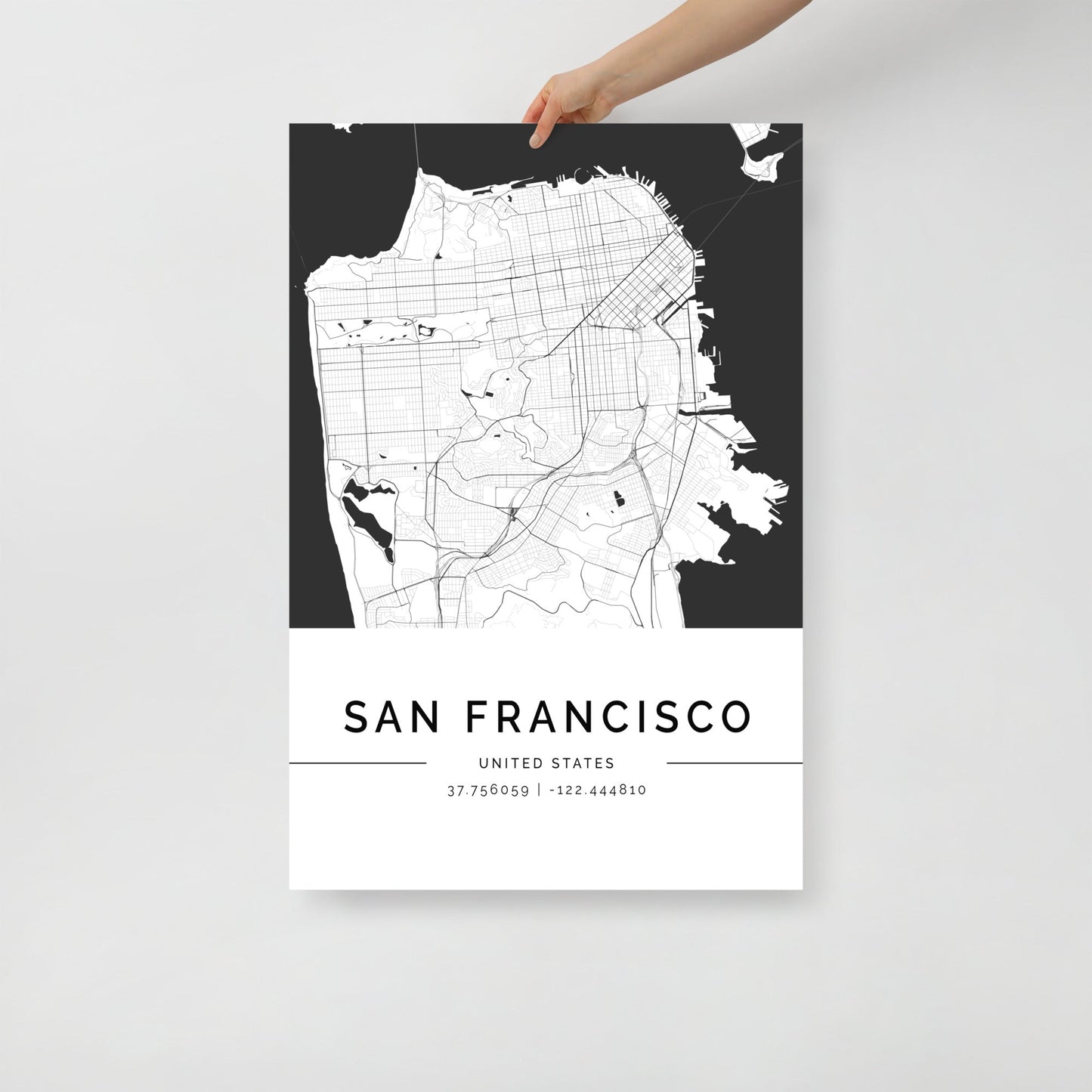 A person holds a black and white San Francisco City Map print by FN Prints, showcasing streets and geographic features ideal for modern interiors. Below, it is inscribed with "San Francisco" and coordinates 37.76959, -122.44810.