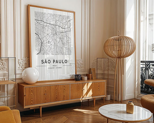 A vintage-style wooden sideboard with decor, including a lamp and sphere, sits beneath FN Prints' São Paulo City Map wall art. This space includes a round coffee table, floor lamp, ornate wall molding, and large windows blending classic charm with modern minimalism.