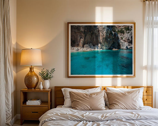 A neatly made bed with white and beige pillows sits by a wooden headboard. Above, FN Prints’ "Sardinian Coast: Part II" framed photo captures the stunning Sardinian cliffs and turquoise sea. A lamp and plant rest on the bedside table.