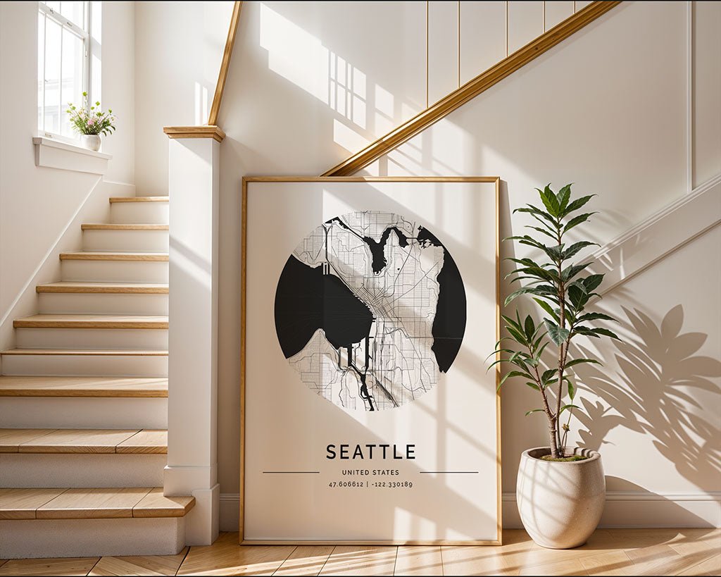 A framed FN Prints Seattle City Map poster rests on the floor at the base of a wooden staircase, showcasing geographic coordinates and urban elegance. Nearby, a potted plant thrives in sunlight streaming through the window, casting shadows on the white wall.