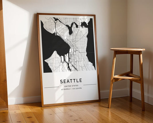 The FN Prints "Seattle City Map," a framed urban landscape print in black and white, leans against the wall on a wooden floor. Nearby, a stool under natural light enhances its appeal as perfect interior decor.