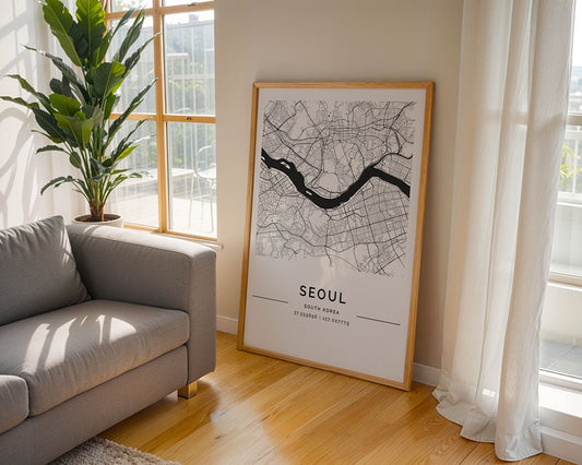 A framed FN Prints Seoul City Map, in black and white with coordinates, leans against the wall in a bright room. A gray sofa complements a tall plant nearby, as sunlight from large windows dances on the wooden floor, capturing the essence of the urban landscape.