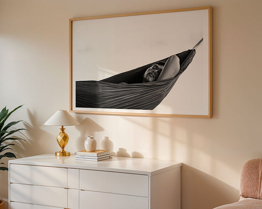 The "Serene Slumber" black and white photograph by FN Prints is framed above a white dresser topped with a gold lamp, plant, and stacked books. Sunlight casts soft shadows, adding tranquil decor to the wall and furniture.