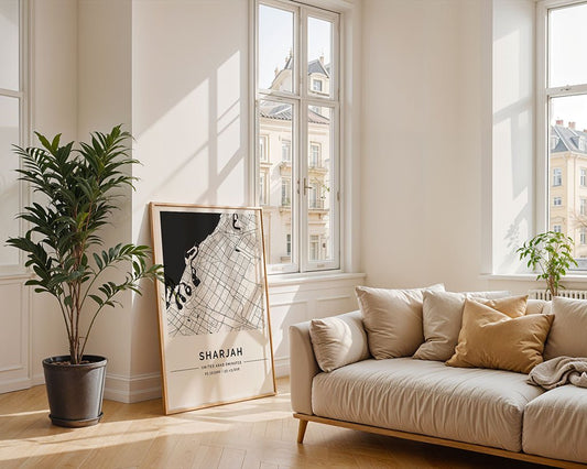 A bright living room features a beige sofa, light pillows, a large potted plant, and the FN Prints Sharjah City Map resting against the wall near tall windows with views of classic buildings.