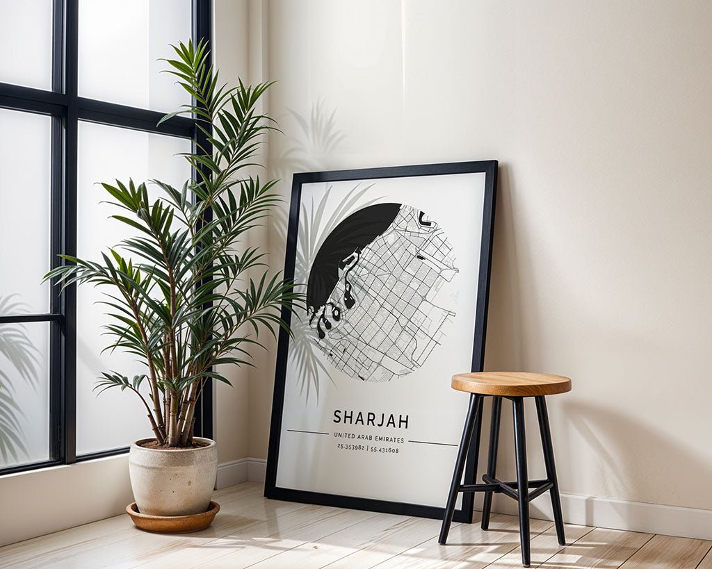 A FN Prints Sharjah City Map, framed and leaning against a white wall, sits beside a potted plant and wooden stool near a large window, with sunlight casting soft shadows on the light floor.