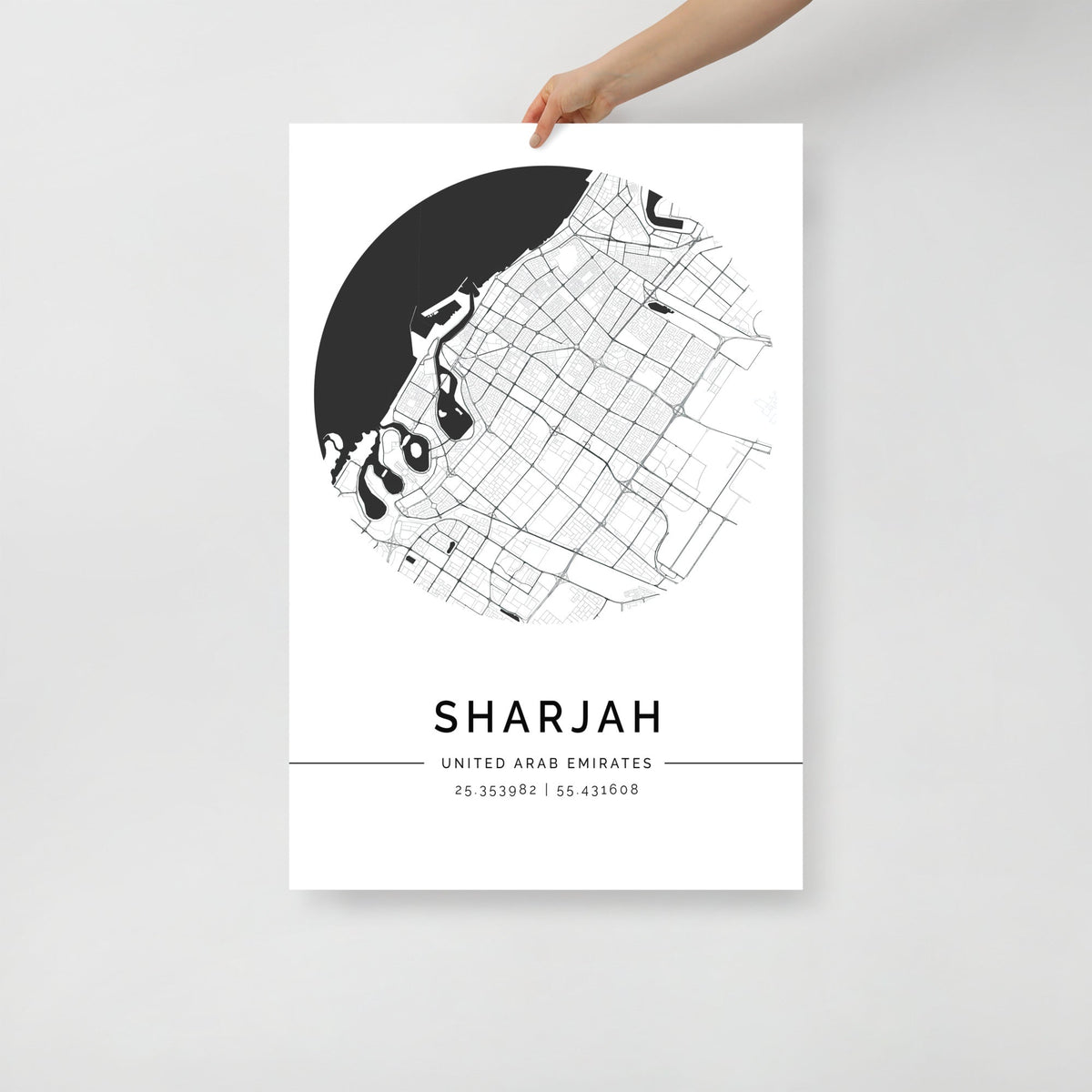 A hand holding a black and white map poster of Sharjah city, with distinct lines representing streets and boundaries on a white background.