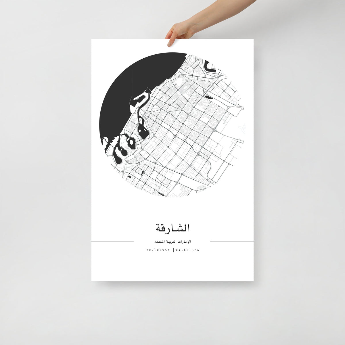 A hand holding a black and white map poster of Sharjah city, with distinct lines representing streets and boundaries on a white background.