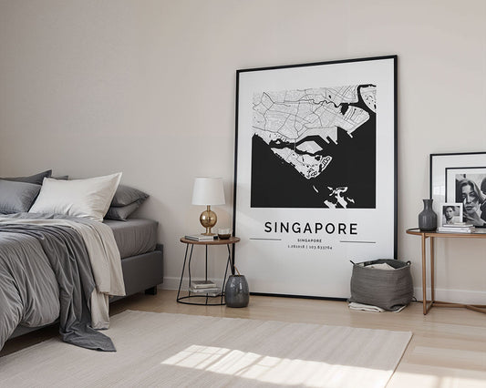 A minimalist bedroom features a large Singapore City Map by FN Prints leaning against the wall. To the left, a bed with gray bedding complements a nightstand decorated with a lamp and various accents. A basket is placed on the floor beneath framed photos on a shelf, adding to the serene atmosphere.