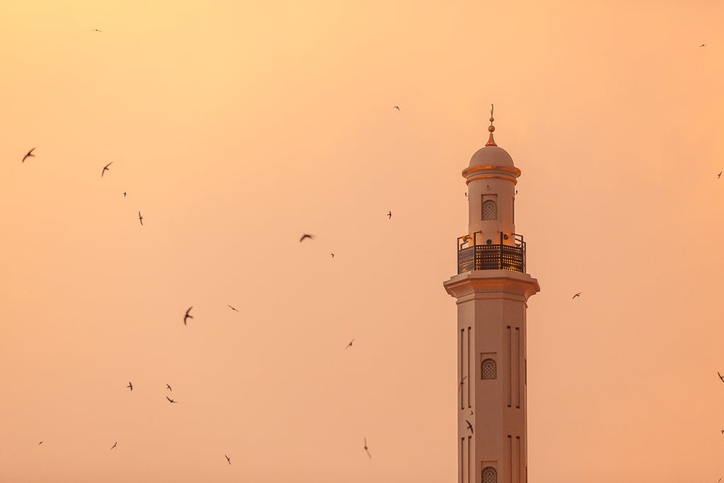 Minaret of Dira mosque in Dubai with birds flying around it at sunset.