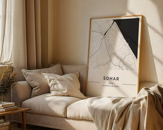 In a cozy living room with a beige sofa and cushions, the FN Prints "Sohar City Map," printed on museum-quality paper with coordinates 24.35915° N 56.71368° E, rests on the sofa. Sunlight highlights plants and a stack of books nearby.