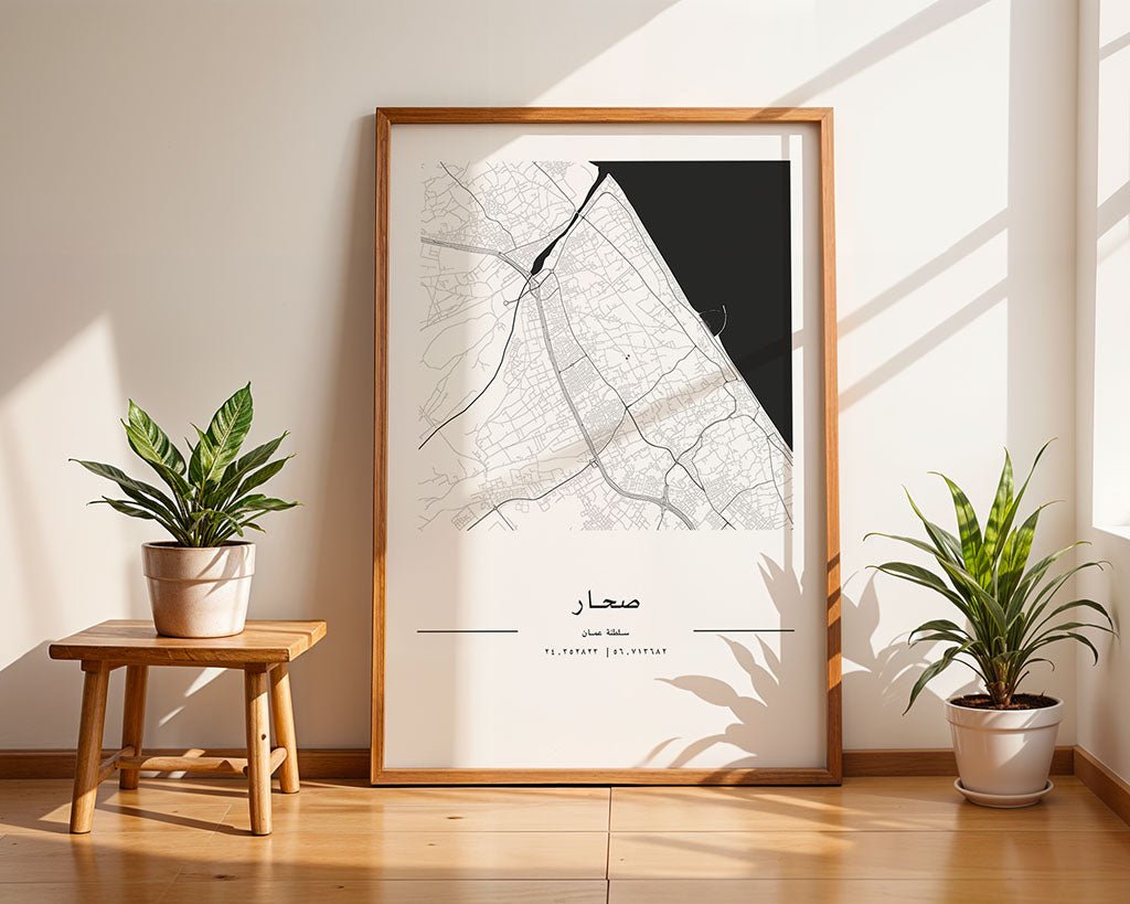 In a well-lit room, a framed FN Prints Sohar City Map with Arabic text leans against the wall. This minimalist print on museum-quality paper depicts a coastal area, complemented by two nearby potted plants enhancing the serene setting.