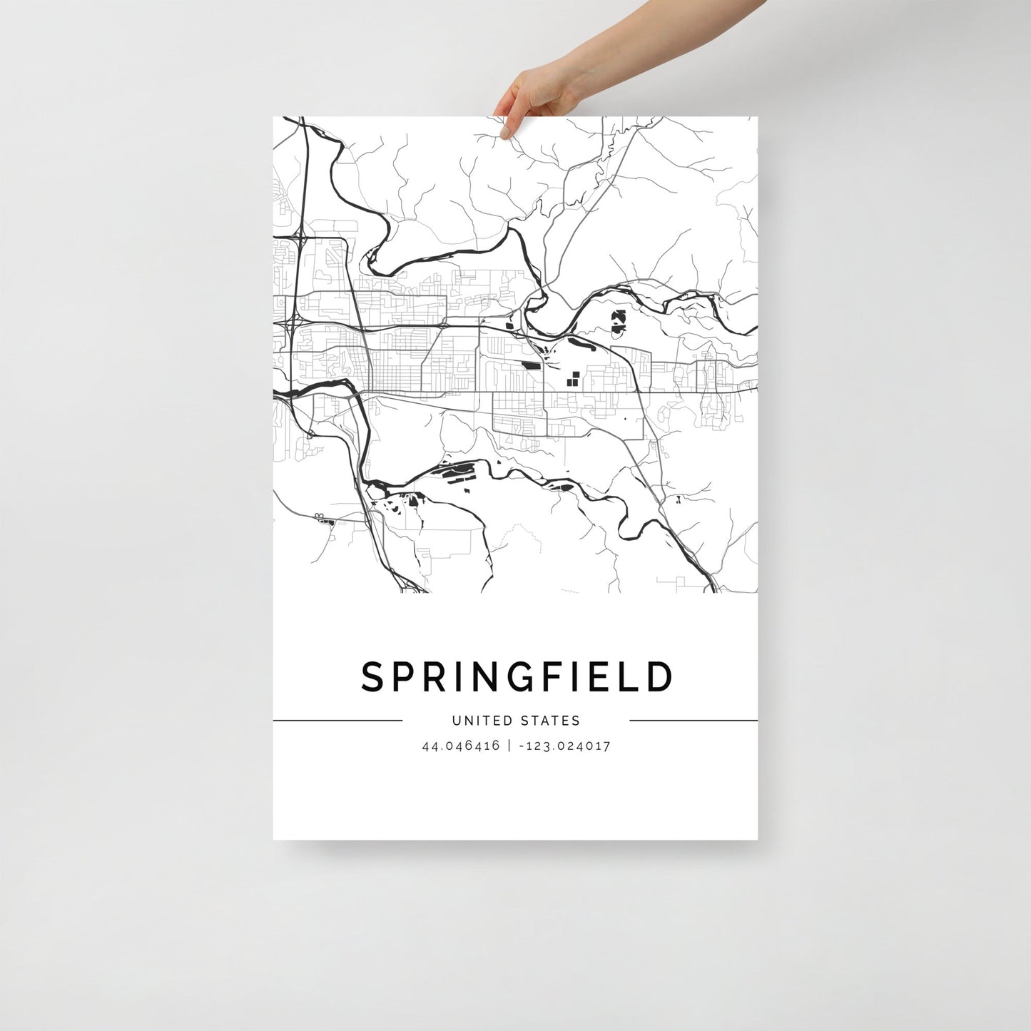 A hand holds a Springfield City Map print by FN Prints, featuring roads and rivers on museum-quality paper. The map's minimalist black-and-white design includes "SPRINGFIELD" and coordinates "44.04616 -123.024017" below.