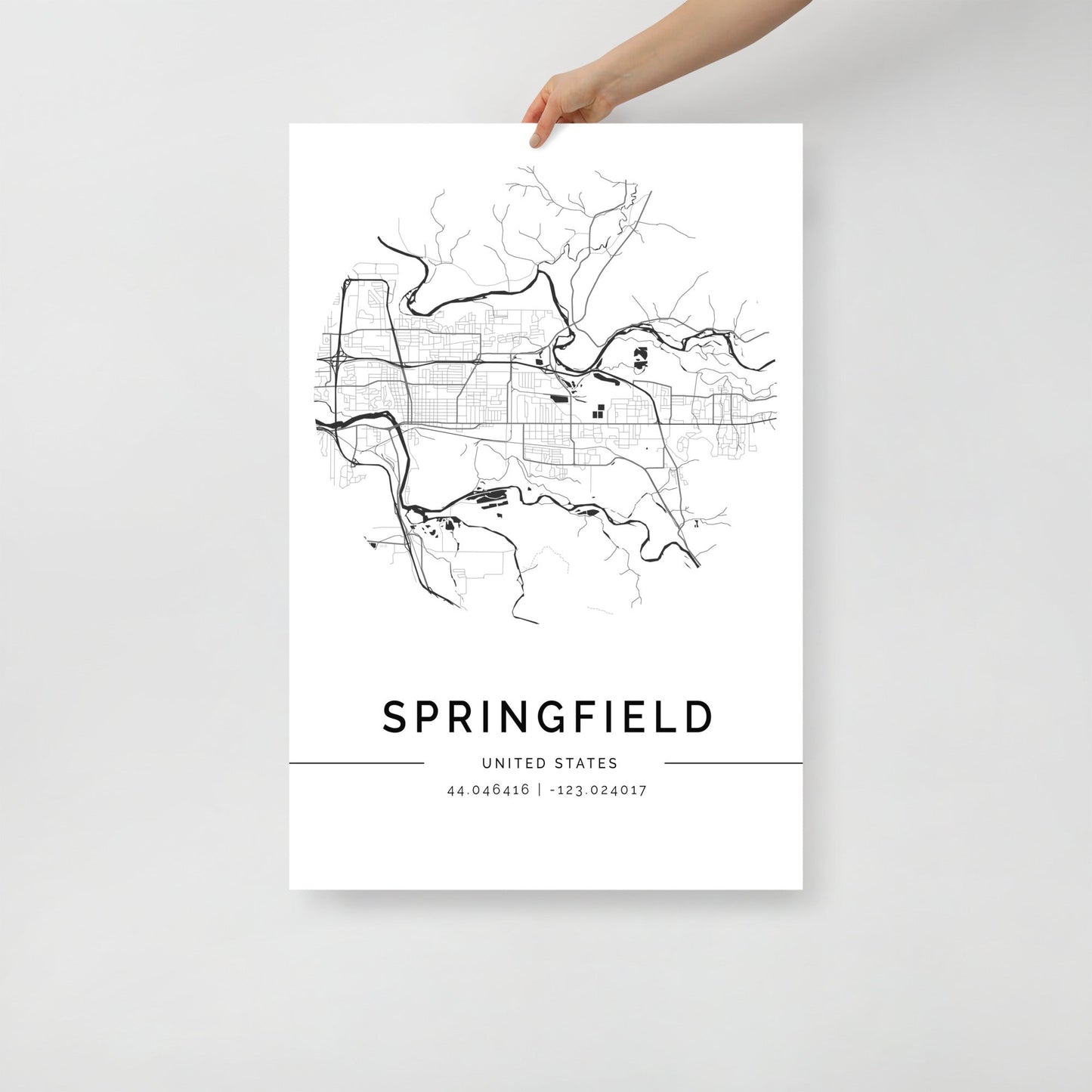 A hand holds an FN Prints Springfield City Map poster, featuring Springfield's street layout in minimalist black and white on fine art paper. Coordinates 44.046416, -123.020417 complement this elegant print.