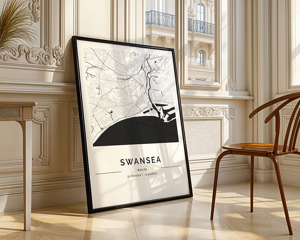 An FN Prints Swansea City Map, elegantly framed on museum-quality fine art paper, leans against a sunlit wall. It meticulously displays streets and coastline coordinates, while a wooden chair and detailed molding complement the minimalist print in this refined setting.