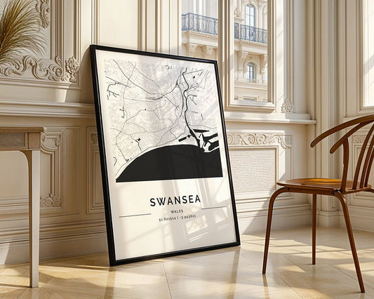 An FN Prints Swansea City Map, elegantly framed on museum-quality fine art paper, leans against a sunlit wall. It meticulously displays streets and coastline coordinates, while a wooden chair and detailed molding complement the minimalist print in this refined setting.