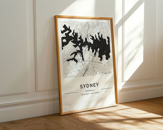 The FN Prints Sydney City Map, with black and white detailing, leans against a white paneled wall. Sunlight illuminates its precise city layout and elegant coordinates on the wooden floor.