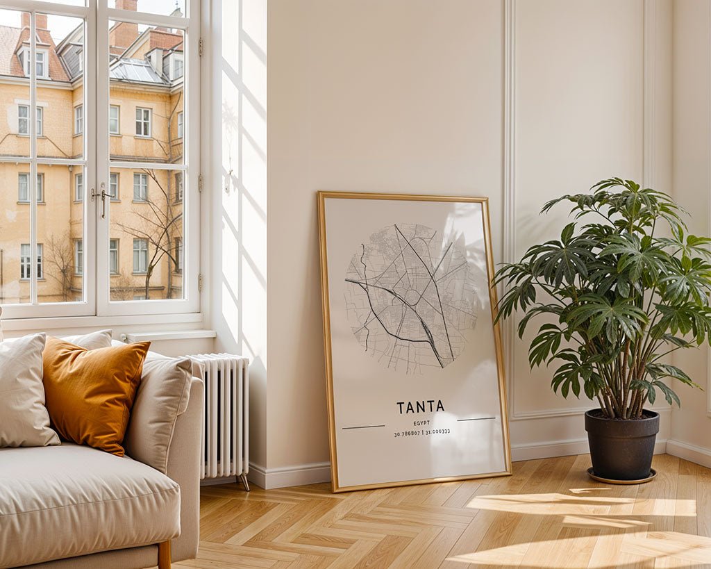 A bright living room with herringbone wood flooring features an FN Prints Tanta City Map leaning against the wall, adding city art charm. A potted plant complements the beige sofa with orange and beige cushions under a window overlooking a building, creating a minimalist aesthetic.