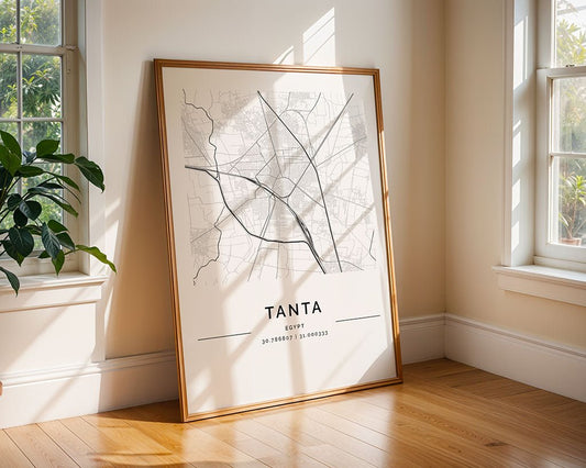 The FN Prints Tanta City Map wall art is framed and leans against a wall in a sunlit, minimalist room with wooden flooring. It highlights major roads and coordinates, positioned near large windows displaying outdoor greenery.