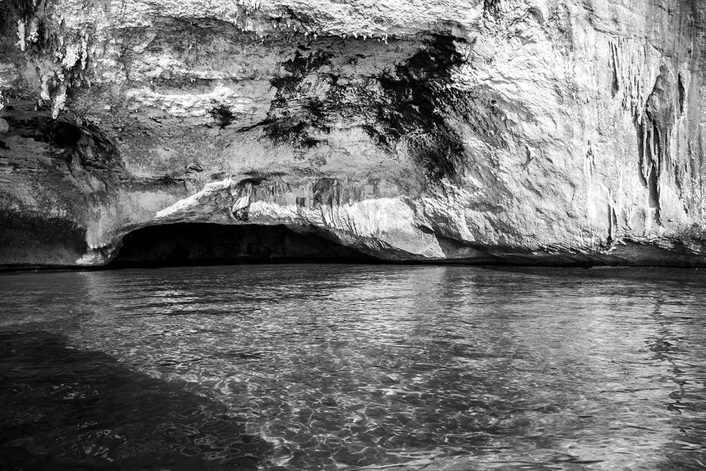The Cave by FN Prints captures a stunning black and white photo of a large rocky cave mouth at the edge of crystal-clear waters. The dark opening of the cave contrasts strikingly with the bright, textured rock surrounding it. Gentle ripples in the clear water reflect light from the rock surface, creating a mesmerizing scene.