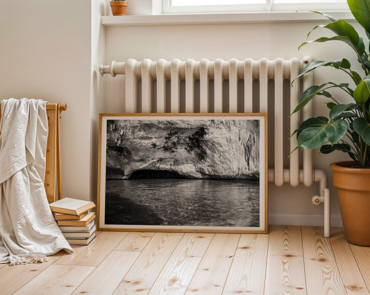 The Cave" by FN Prints, a framed black and white coastal cliff photograph, leans against a white radiator. Nearby are a terracotta potted plant, stacked books, and a draped blanket on the light wood floor.