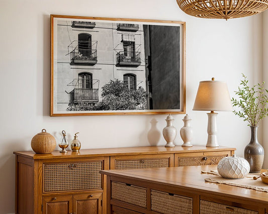 A dining room with a wooden table and sideboard, adorned with vases, a lamp, woven baskets, a plant, and wall decor featuring "The Reader," a large framed black-and-white FN Prints photo of balconies.
