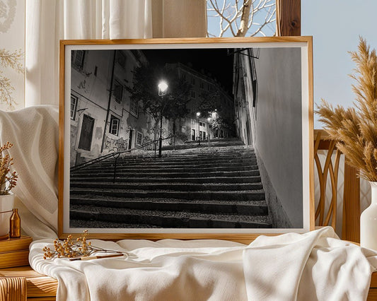 The Staircase by FN Prints, a framed black and white Lisbon staircase photograph, is displayed on a draped cloth in a cozy room with soft lighting, dried plants, and tall curtains—perfect urban wall art.