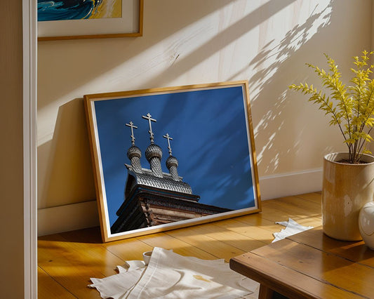 The "Three Crosses" framed photo by FN Prints, showcasing a church roof with three domes and crosses, leans against a beige wall in a sunlit room with a potted plant and white rug on wooden floors.