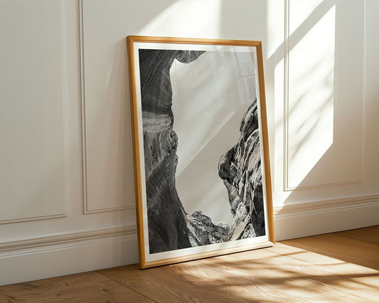 Through The Ravine by FN Prints is a framed black and white art print featuring a rocky canyon, displayed against a white paneled wall on a wooden floor, sunlight casting shadows—an elegant piece of minimalist landscape photography.