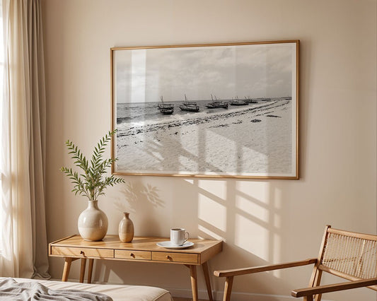 A framed "Tourist Boats At Nakupenda Sandbank" black-and-white photo by FN Prints hangs above a wooden console with two vases, a cup and saucer, and a rattan chair. Sunlight filters through sheer curtains on the beige wall.