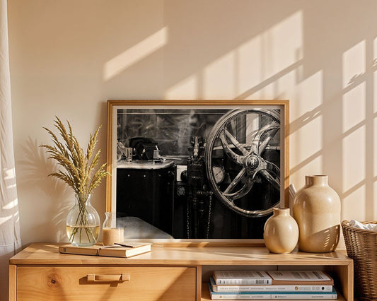 A framed black-and-white “Tram Cab” photograph by FN Prints sits on a wooden sideboard, alongside a clear vase with dried grasses, two beige ceramic vases, and a stack of books. Sunlight casts soft shadows across the wall.