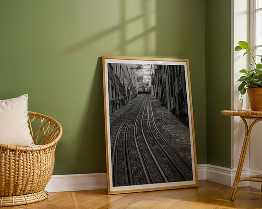 Tram Tracks" by FN Prints, a black-and-white framed tram wall art photograph, leans against a green wall in a sunlit room featuring wooden floors, a rattan chair with cushion, and a small table topped with a potted plant.