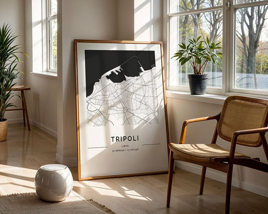 A framed Tripoli City Map by FN Prints leans against a sunlit wall, featuring coordinates at the bottom. Nearby are a wooden chair, white stool, potted plants, and large windows illuminating the minimalist black and white design with natural light.