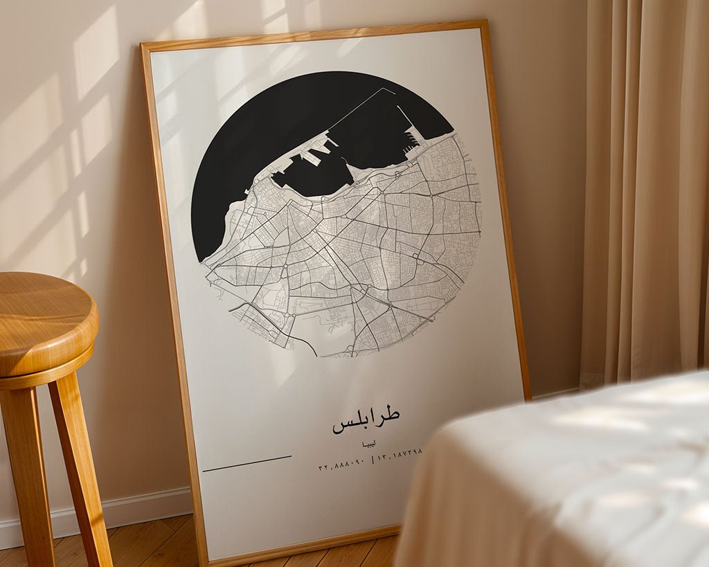 A framed FN Prints Tripoli City Map poster, showcasing intricate street lines in a black circular design, leans against a beige wall in a softly lit room. A wooden stool sits on the left, with beige curtains and a bed partially framing the minimalist scene.