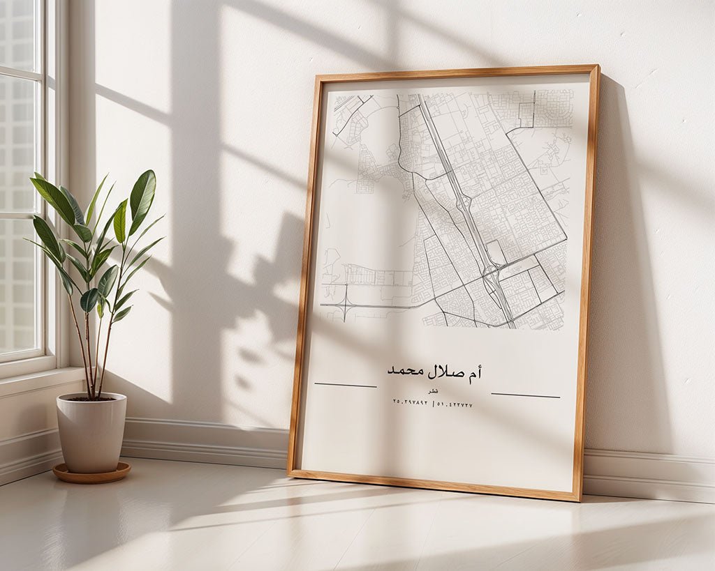 A framed FN Prints Umm Salal Mohammed City Map leans against a sunlit white wall, with Arabic text visible. A potted plant sits nearby on the wooden floor, while windowpane shadows cross the wall and floor.