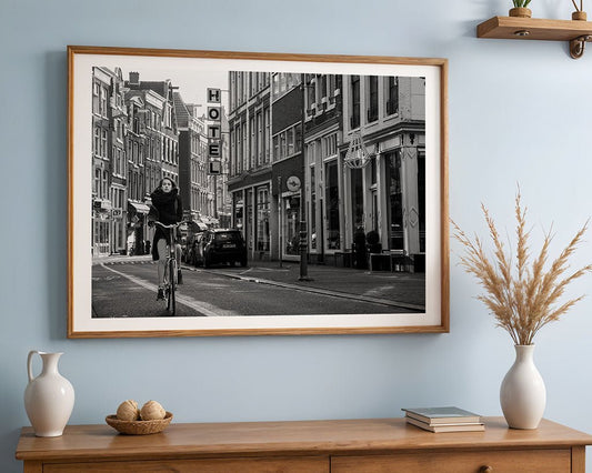The FN Prints "Urban Cyclist" framed black and white Amsterdam photo shows a biker on a city street, hanging above a wooden shelf with decor—ideal urban wall art for modern spaces.