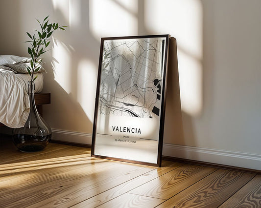 The FN Prints Valencia City Map poster, with its minimalist design, rests on a wooden floor by the bed. Sunlight casts shadows on the wall, enhancing the sophisticated décor. A nearby glass vase holds a slender plant, adding elegance to this serene setting.