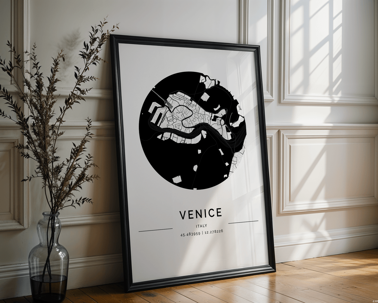 A framed black and white FN Prints Venice City Map with coordinates 45.439589 | 12.331226 leans against a sunlit white paneled wall, casting shadows on the floor, while a clear vase with branches complements the minimalist style.