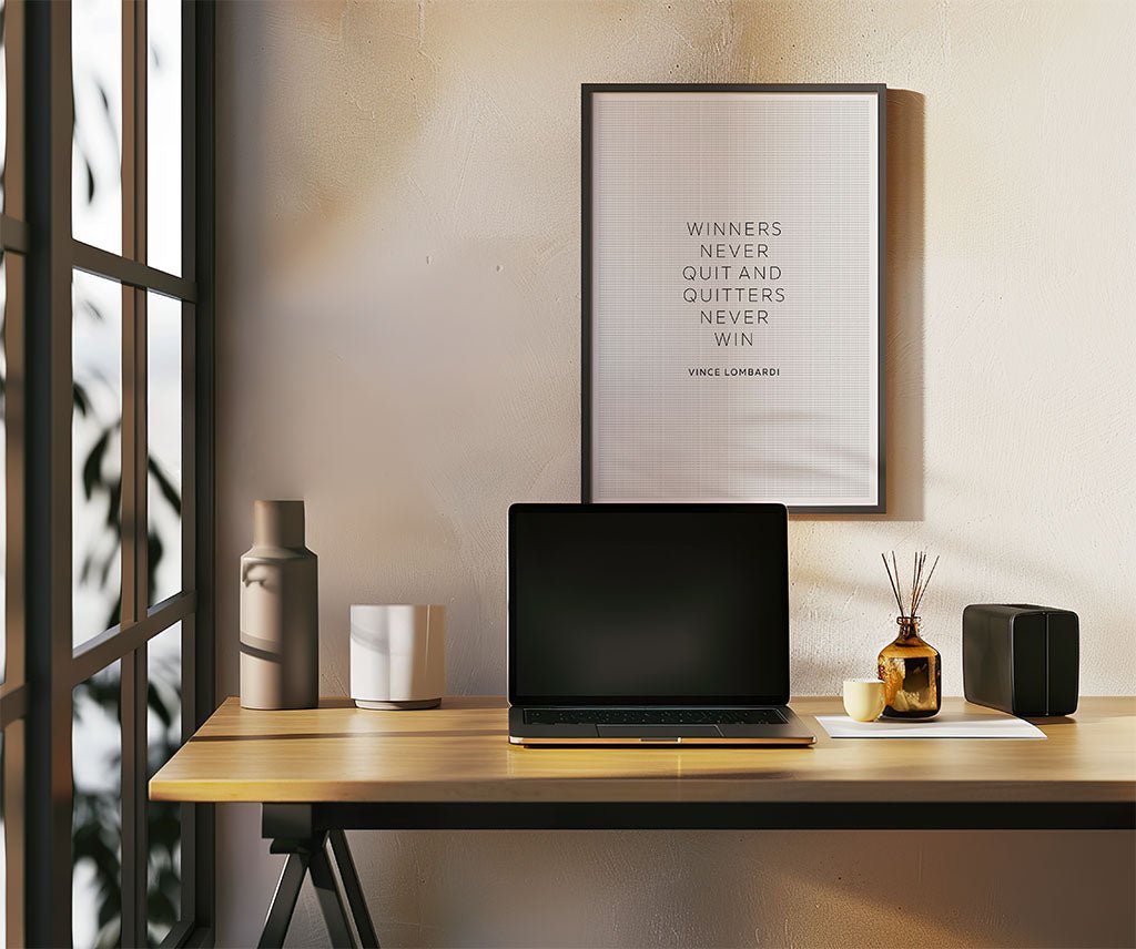 A minimalist desk features a closed laptop, small vases, a notebook, and the FN Prints Vince Lombardi - Winners Never Quit wall art—a motivational print with the quote, “Winners never quit and quitters never win.”.