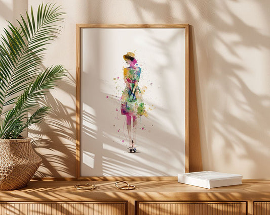 The FN Prints "Watercolour Woman" framed wall art features a woman in a colorful dress and hat, displayed on wood beside a potted plant, jewelry, and book. Leafy shadows enhance this modern abstract decor piece.