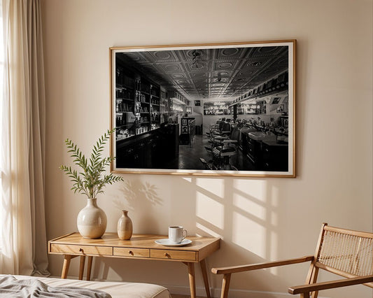 The "Welcome To The Baba's House" framed print by FN Prints hangs above a wooden table with a vase, plant, and cup. Sunlight filters in through a curtained window, casting soft shadows and enhancing this charming retro barber shop decor.